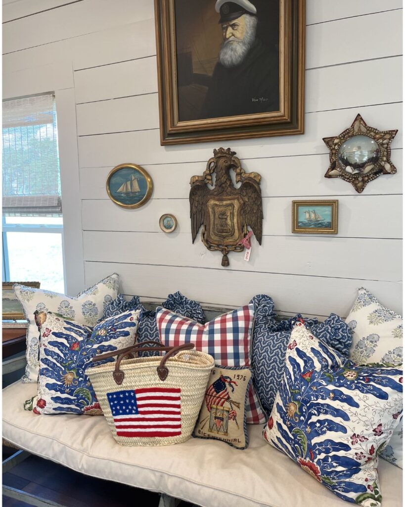 A cozy bench with blue and white floral pillows, a plaid pillow, and bags with American flag designs. Nautical-themed artwork by an interior designer Boerne TX hangs on the white shiplap wall above.