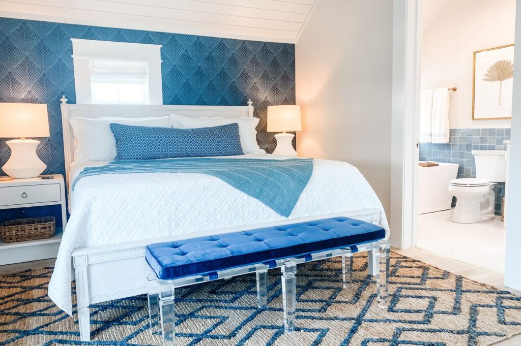 A bright bedroom by an interior designer in Boerne, TX, featuring a white bed with blue and white bedding, blue accent wall, white nightstands with lamps, blue bench, and a view into a bathroom with blue tiles.