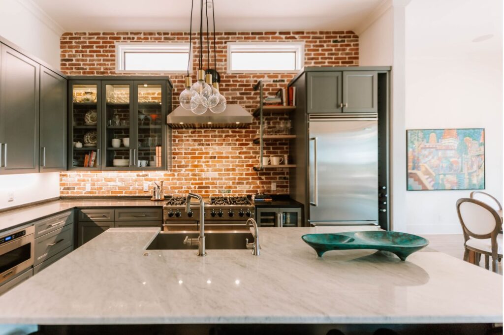 Modern kitchen with a marble island, double sink, green decorative bowl, stainless steel appliances, dark cabinets, and exposed brick wall—expertly styled by an interior designer Boerne TX—with high windows and pendant lights above the island.