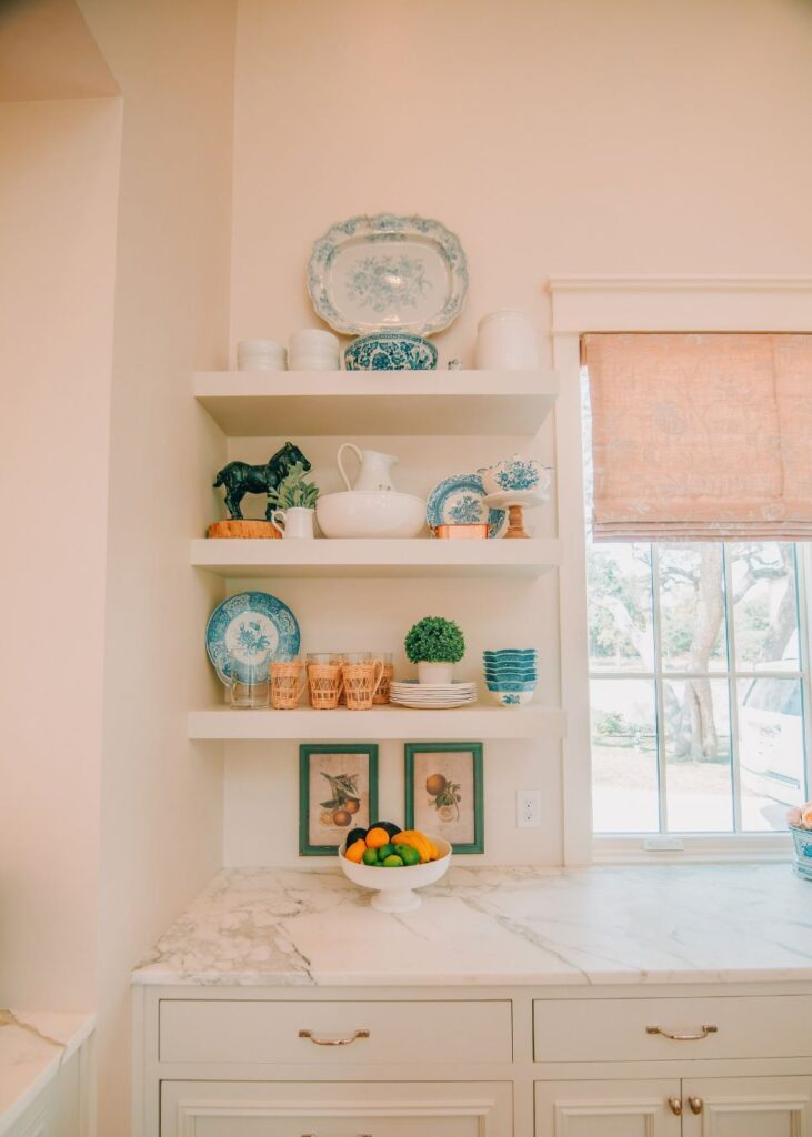Open shelves in a bright kitchen by an interior decorator Boerne display blue and white dishes, cups, and decor. Framed art prints hang above a marble countertop with fresh citrus, while a large window brings in natural light.