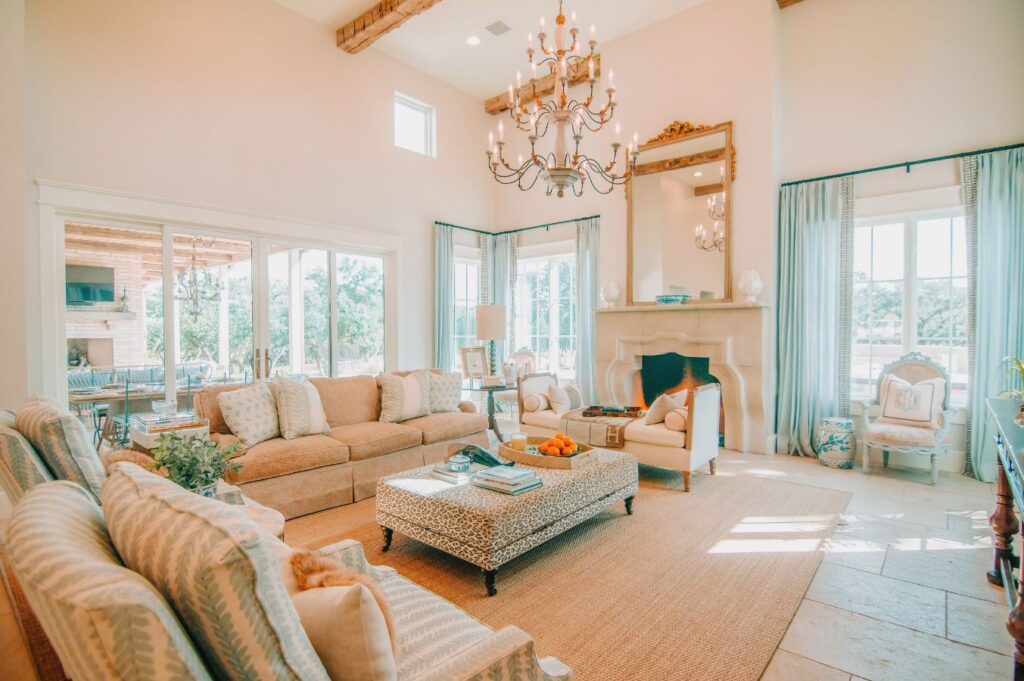Bright, elegant living room designed by an interior decorator Boerne, with beige sofas, a patterned ottoman coffee table, a fireplace topped by a large mirror, chandelier, light blue curtains, and large windows streaming natural light.