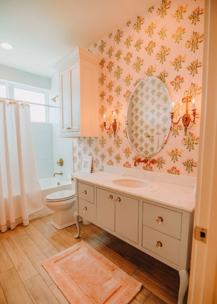 A bright bathroom by an interior designer in Boerne, TX, featuring floral wallpaper, an oval mirror, white vanity, gold fixtures, a white shower curtain, pink rug on wood-look flooring, and wall sconces flanking the mirror.