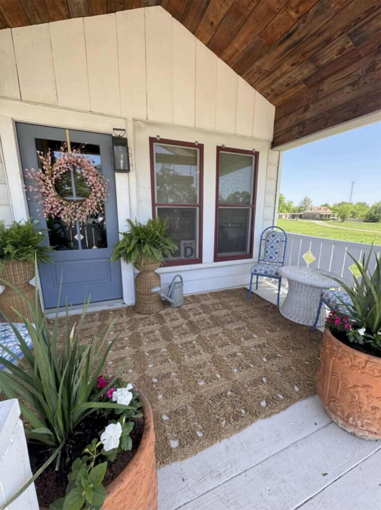 A cozy farmhouse porch styled by an interior decorator Boerne, with a blue door adorned with a pink wreath, potted plants, a blue chair, watering can, and woven rug. Large windows overlook the grassy yard and open landscape.