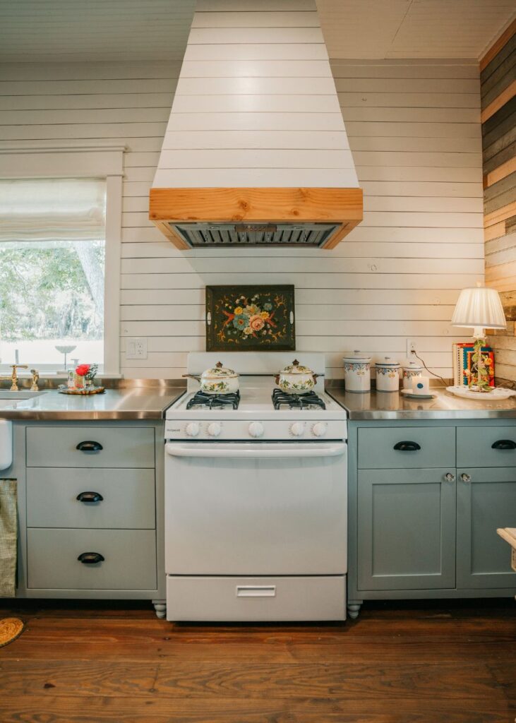 A cozy kitchen with light blue cabinets, a white gas stove, and stainless steel countertops—styled by an interior designer Boerne TX—with canisters and a lamp on the right counter and a window on the left.