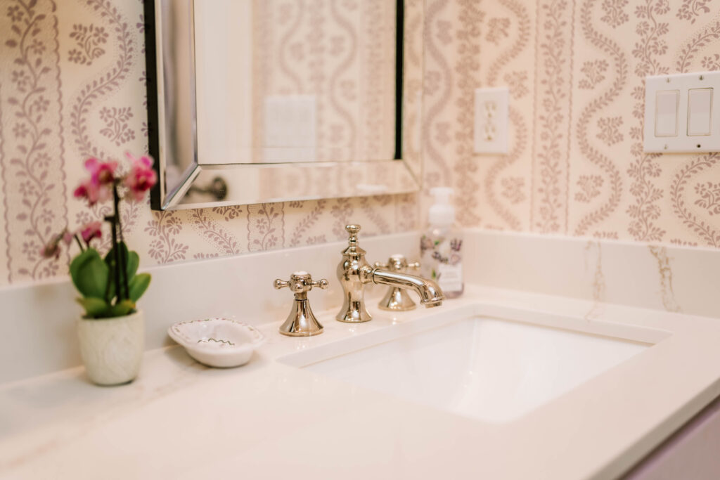 A bathroom sink with gold faucet, white countertop, and ornate wallpaper styled by an interior decorator Boerne. A small pink flower in a pot, soap dispenser, and dish with jewelry sit beside the sink. A mirror is mounted above.