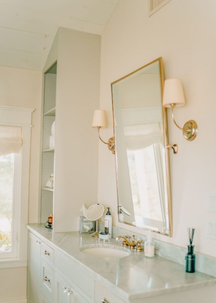 A bright bathroom vanity with a marble countertop, gold fixtures, and a large mirror showcases the elegant touch of an interior design company in Boerne, TX—perfectly arranged shelves and toiletries complete the sophisticated look.