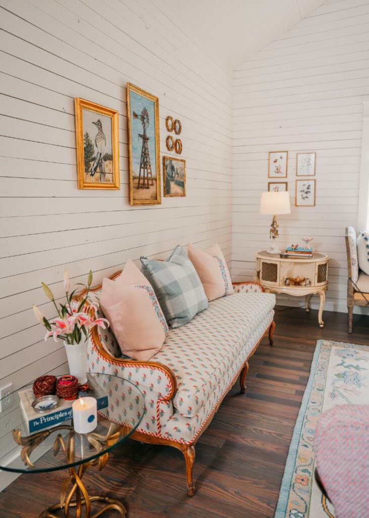 A cozy living room styled by an interior designer Boerne TX, featuring a vintage sofa with pastel cushions, a side table with flowers and candle, framed art on white shiplap walls, and a small lamp on a side cabinet.