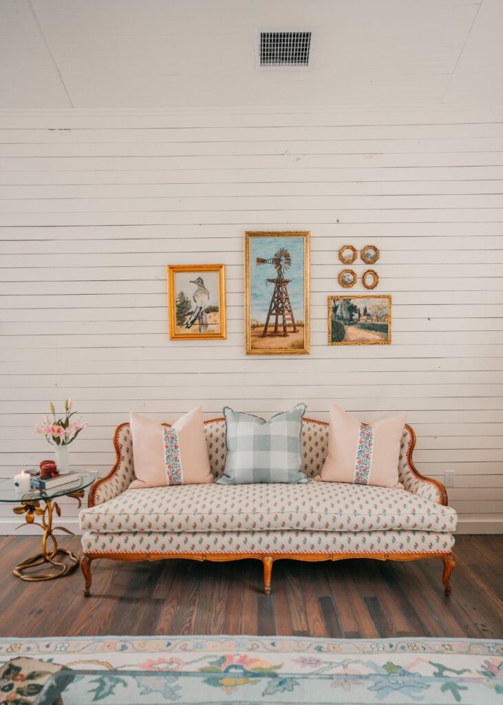 A vintage sofa with patterned upholstery and pastel pillows sits against a white shiplap wall, styled by an interior designer Boerne TX, with gold-framed artwork and a small round side table holding a vase of flowers.
