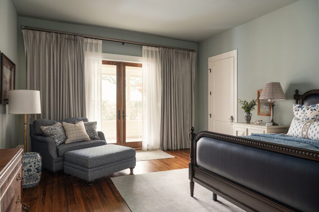 A cozy bedroom by an interior decorator Boerne features a dark wood bed, blue and white bedding, a chaise lounge with pillows, hardwood floors, a white rug, dresser, and large windows with sheer and gray curtains letting in natural light.