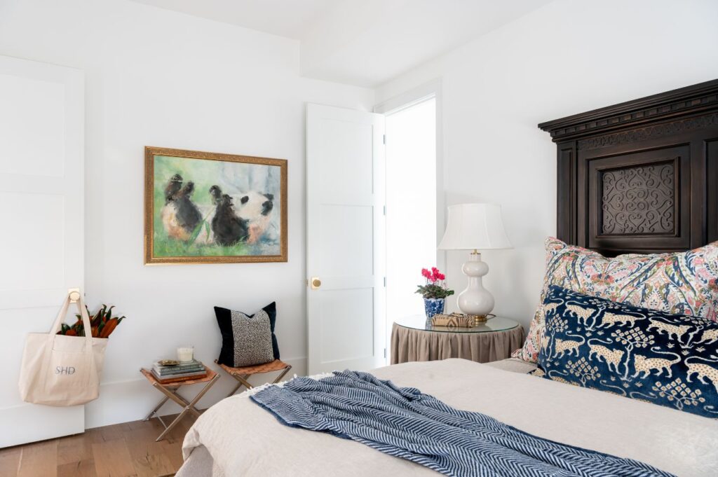 Bright bedroom with white walls, a dark wooden headboard, patterned pillows, and vibrant details styled by an interior designer Boerne TX—featuring a chic side table, pink flowers, playful animal painting, and a tote bag hanging on the door.