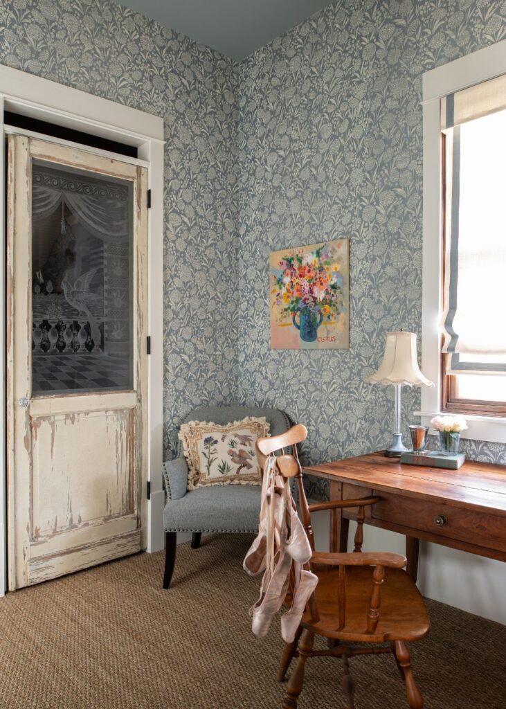 A cozy room with blue floral wallpaper, vintage door, light blue upholstered chair, wooden desk with a lamp, window, ballet slippers on a chair, and vibrant flower painting—perfect inspiration from an interior decorator Boerne would admire.