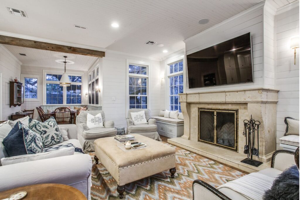 A cozy living room, styled by an interior designer Boerne TX, features white walls, a beige stone fireplace, a mounted TV, cushioned seating, ottoman, patterned rug, and large windows. A dining area is visible in the background.