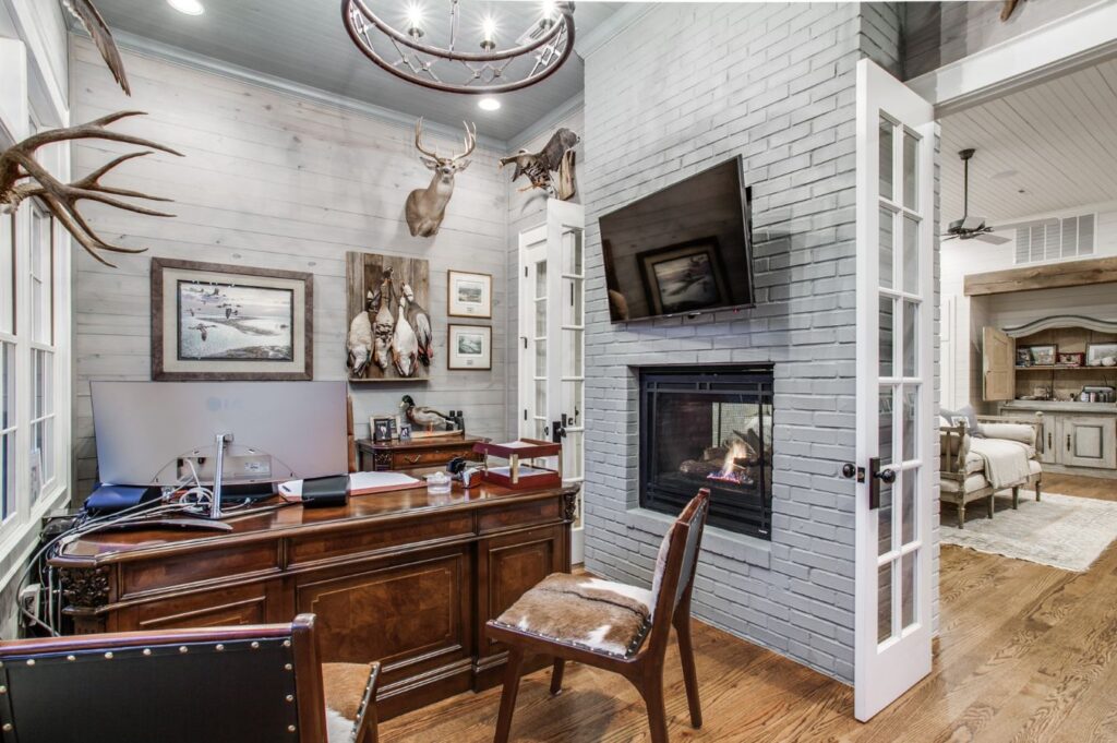 A rustic home office designed by an expert interior designer in Boerne, TX, features a wooden desk, cowhide chair, mounted deer heads, animal photos, a gray brick fireplace, and a wall-mounted TV leading into a cozy living room.