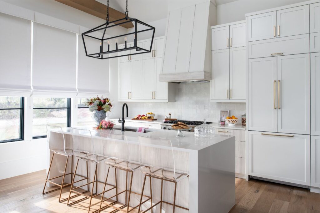 Modern white kitchen with a large island featuring a glossy countertop, four gold and white bar stools, black faucet, bouquet of flowers, pendant light, and floor-to-ceiling cabinets. Perfect inspiration for interior design Boerne TX enthusiasts. Large windows provide natural light.