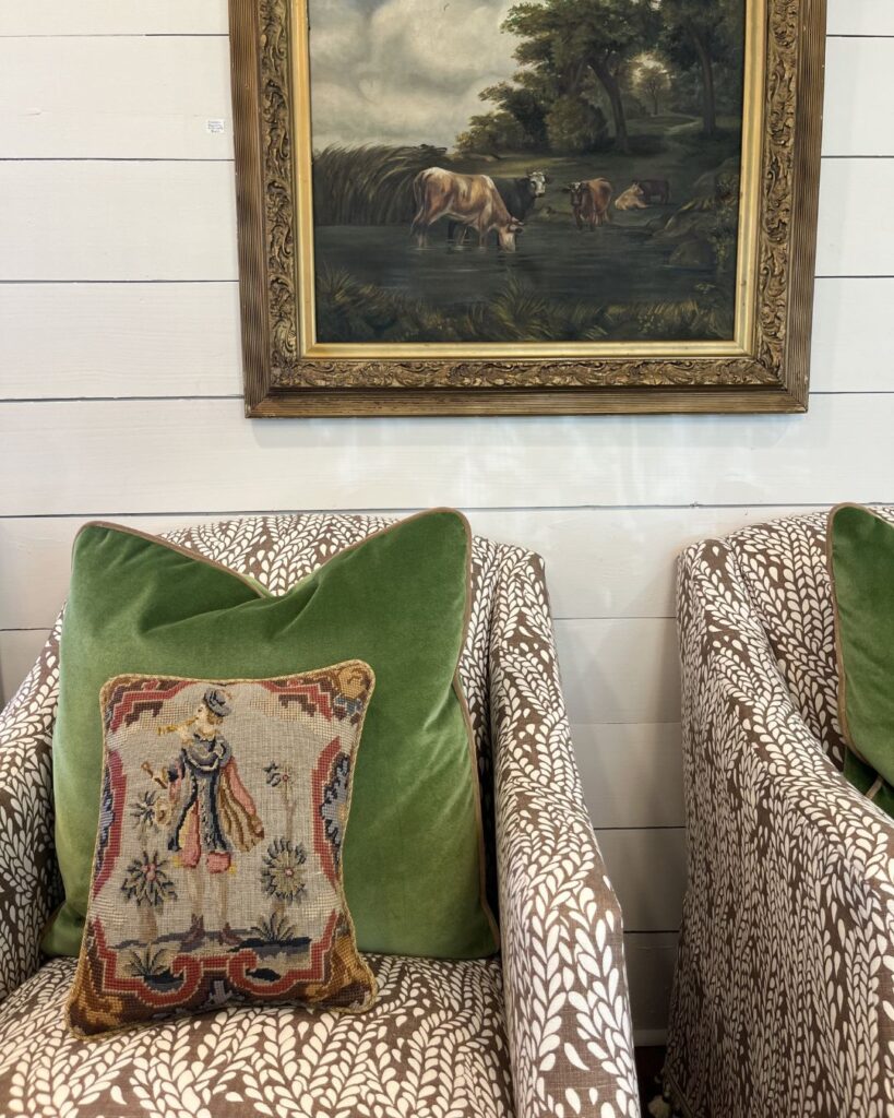 Two patterned armchairs with green velvet pillows sit side by side; one has an embroidered cushion featuring a figure. Above, a framed painting of cows by water hangs on a white shiplap wall—an inviting touch from an interior designer Boerne TX.