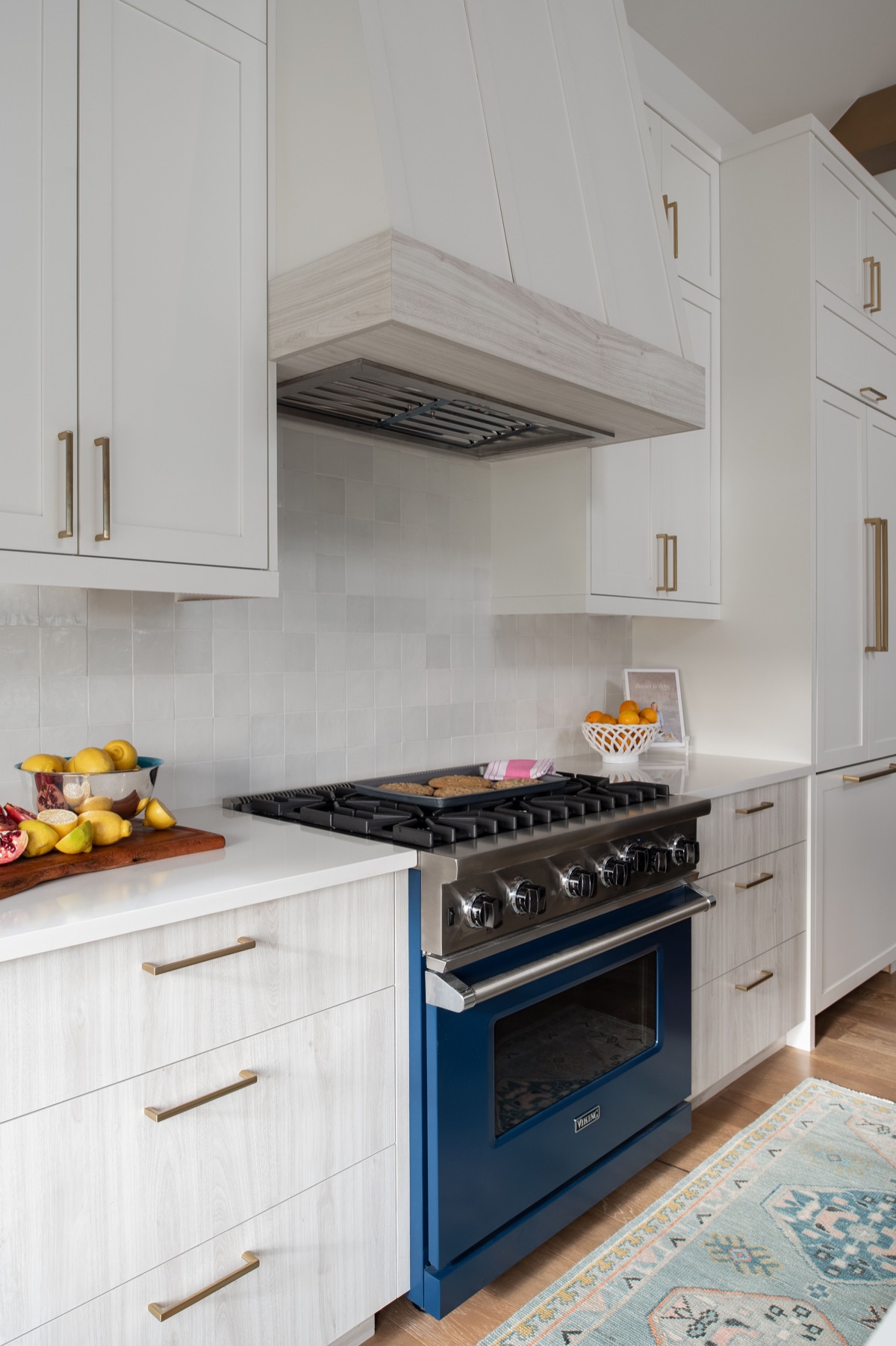 A deep blue enamel range with a stainless steel cooktop under a pale wood and shiplap hood, set into pale wood cabinetry with brass bar pulls and a white zellige backsplash, with lemons and pomegranates on a wooden board.