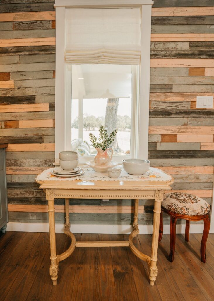 A small vintage wooden table with dishes and a flower vase sits beneath a window, against a rustic wooden plank wall. Styled by an interior decorator Boerne, a floral stool is beside the table on the wooden floor.