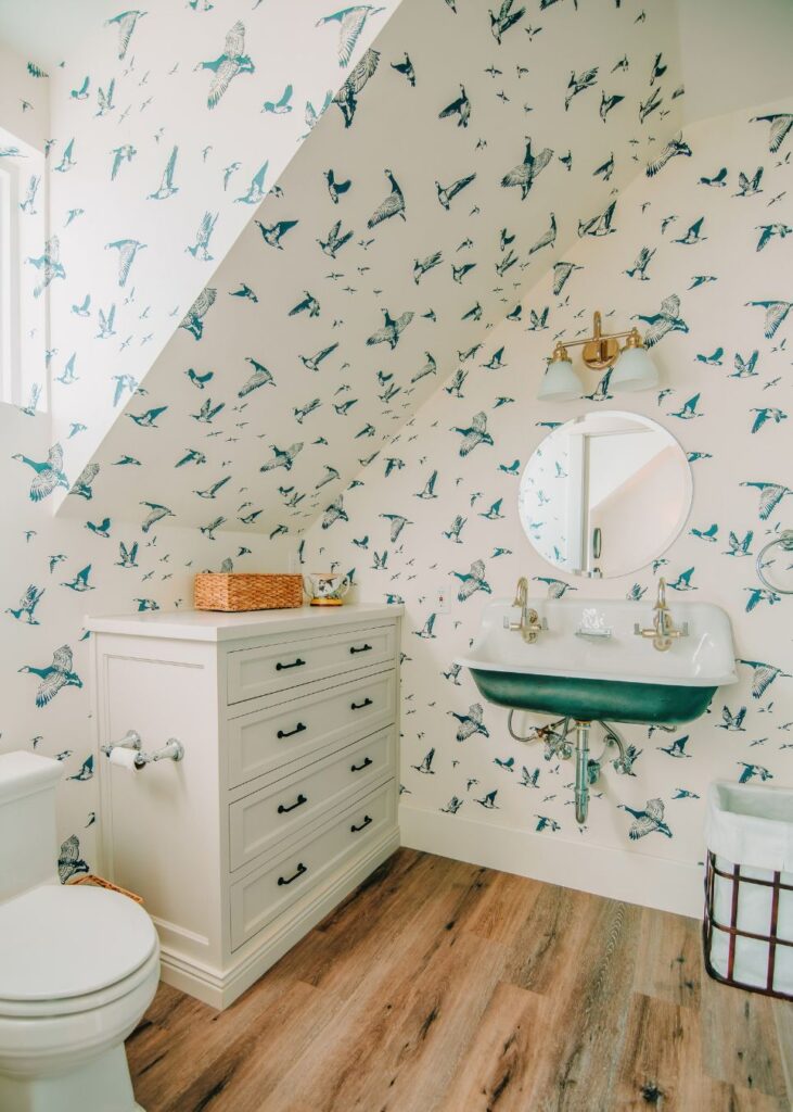 A bright bathroom with bird-patterned wallpaper, a round mirror above a wall-mounted sink, white vanity drawers, a toilet, and wood-style flooring under a sloped ceiling designed by an interior designer Boerne TX.
