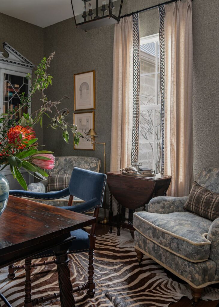 A cozy sitting room, curated by an interior decorator in Boerne, features two patterned armchairs with plaid cushions, a dark wood table, animal print rug, framed art, and tall curtains framing a window aglow with soft light.