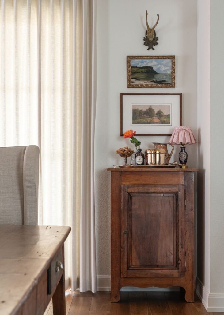 A wooden cabinet topped with a lamp, flowers, and bottles stands against a light wall. Above it are two framed landscape paintings and a small decorative antler mount. Sunlight filters through sheer curtains to the left.