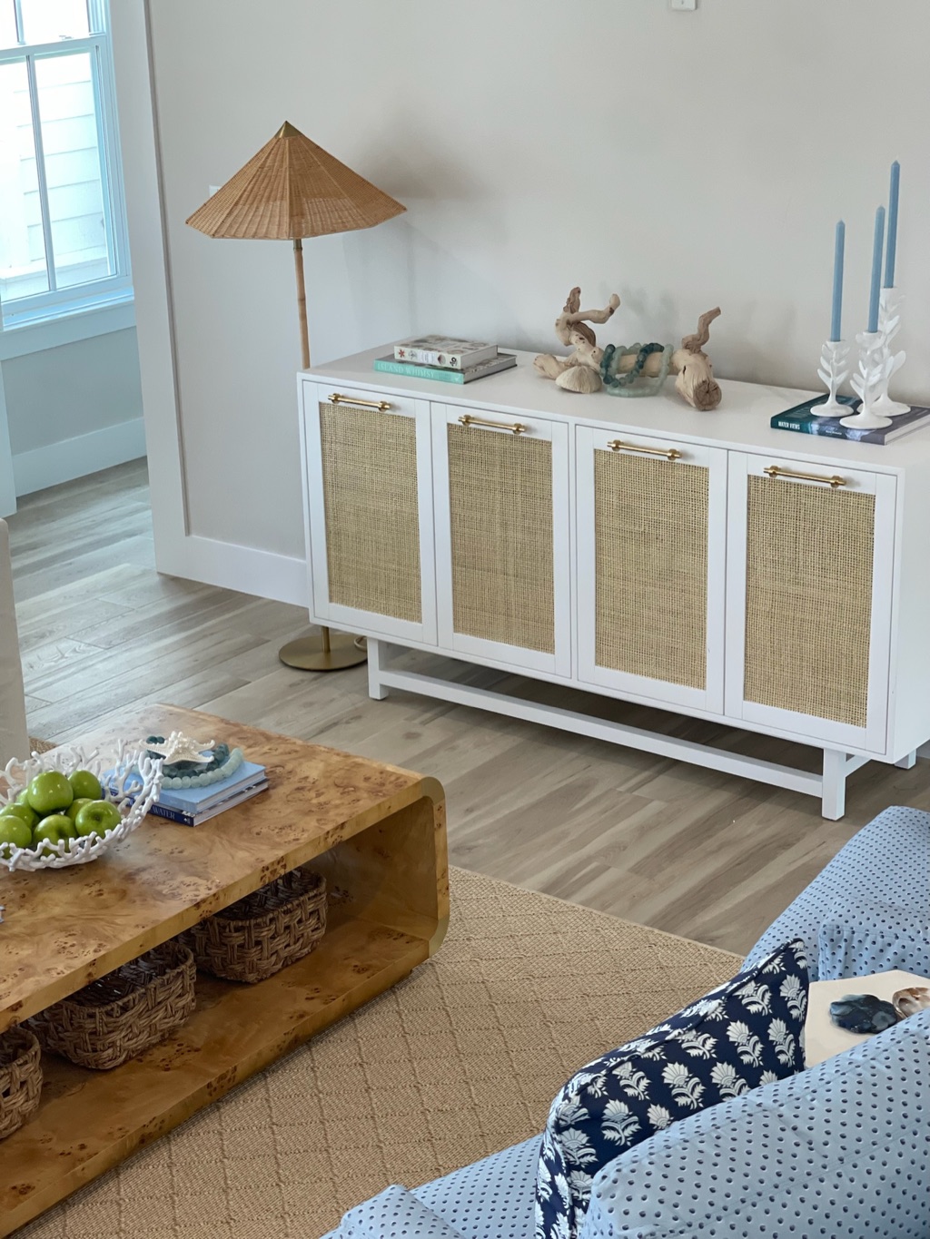 A white console cabinet with woven cane door fronts and slim brass pulls, topped with driftwood, coral books and pale blue candlesticks, beside a rattan shade floor lamp, with a burl wood coffee table and a seagrass rug in the foreground.