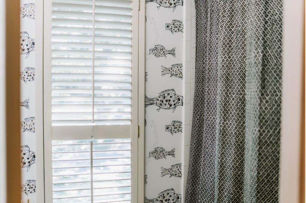 A window with white shutters is partially open. The wall is adorned with fish-themed wallpaper, and a bold black-and-white curtain hangs to the right—a charming touch from an interior designer in Boerne, TX.
