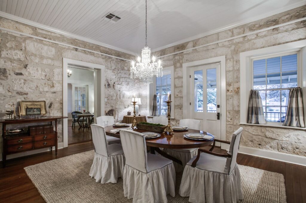Elegant dining room with stone walls, a round wooden table set for four, white slipcovered chairs, a chandelier, and large windows. Styled by an interior designer Boerne TX, the space features hardwood floors and rustic, vintage decor.