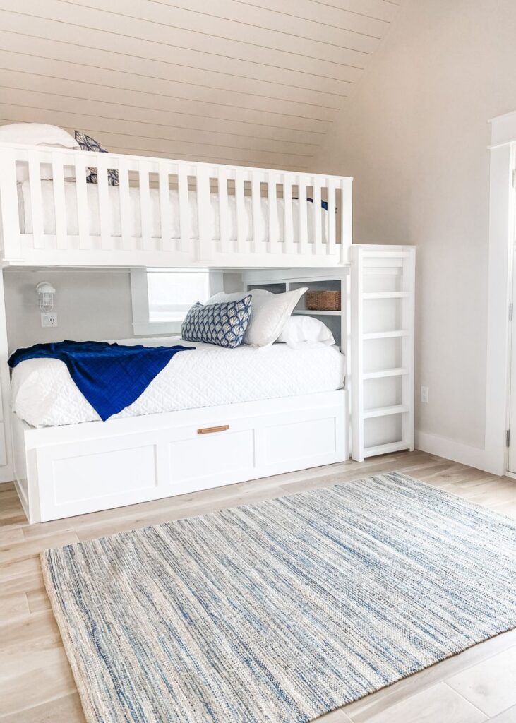 White wooden bunk beds with white bedding and blue accent pillows, a blue throw blanket, and built-in drawers beneath the lower bunk. A light blue striped rug covers the wooden floor in this bright, airy space by an interior design company Boerne TX.