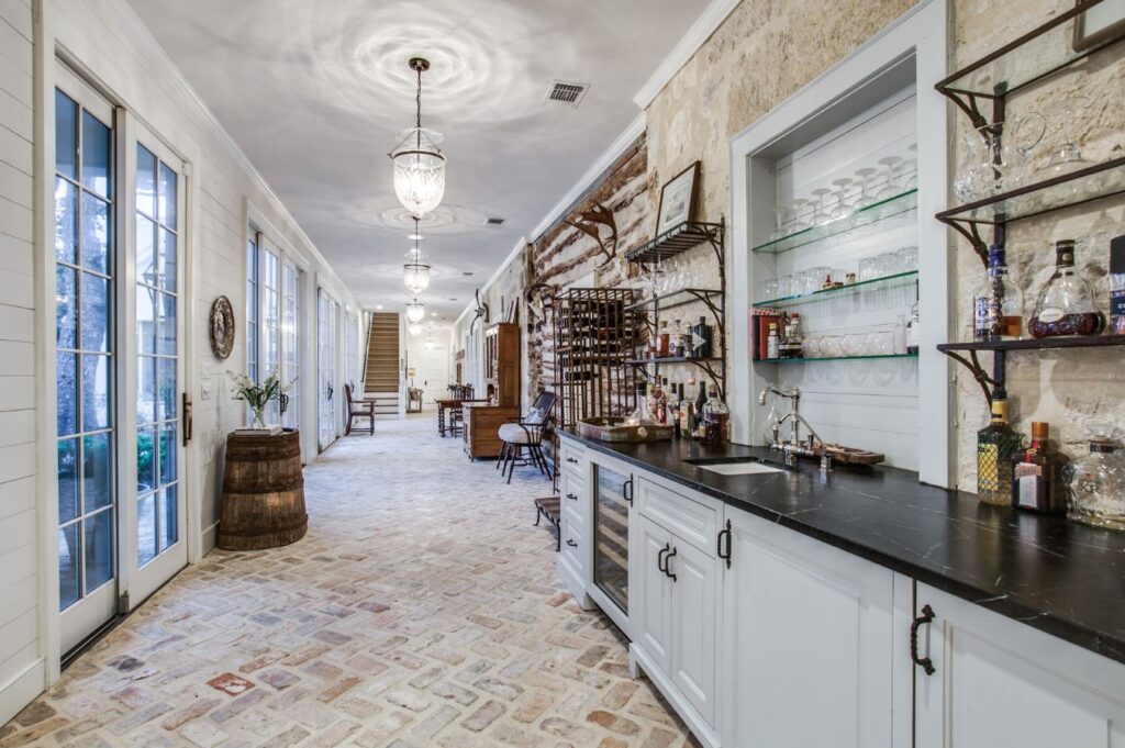 A long, elegant hallway with exposed brick flooring, glass doors on one side, and a bar area with glass shelves, drinks, and a sink—perfect inspiration for anyone seeking interior design in Boerne TX. Rustic decor and vintage furniture line the corridor.