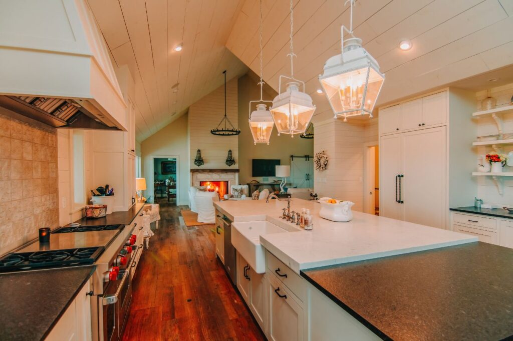 A spacious, modern farmhouse kitchen with white cabinets, pendant lights, a large island, dark countertops, and wooden floors—expertly crafted by an interior designer Boerne TX—flows into a cozy living area with a fireplace.