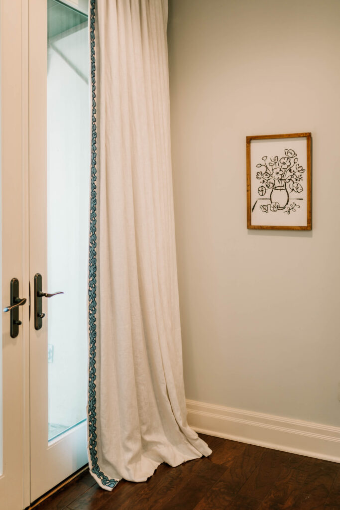 A white curtain with a blue-trimmed edge hangs by glass double doors. Styled by an interior designer in Boerne, TX, a framed black-and-white line drawing of flowers decorates the light wall above a warm wooden floor.