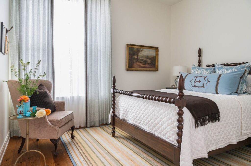 A cozy bedroom by an interior design company in Boerne, TX, featuring a dark wood bed, white quilt, blue pillows, a striped rug, and taupe armchair beside a small table with flowers. Light pours in through sheer curtains at the tall window.