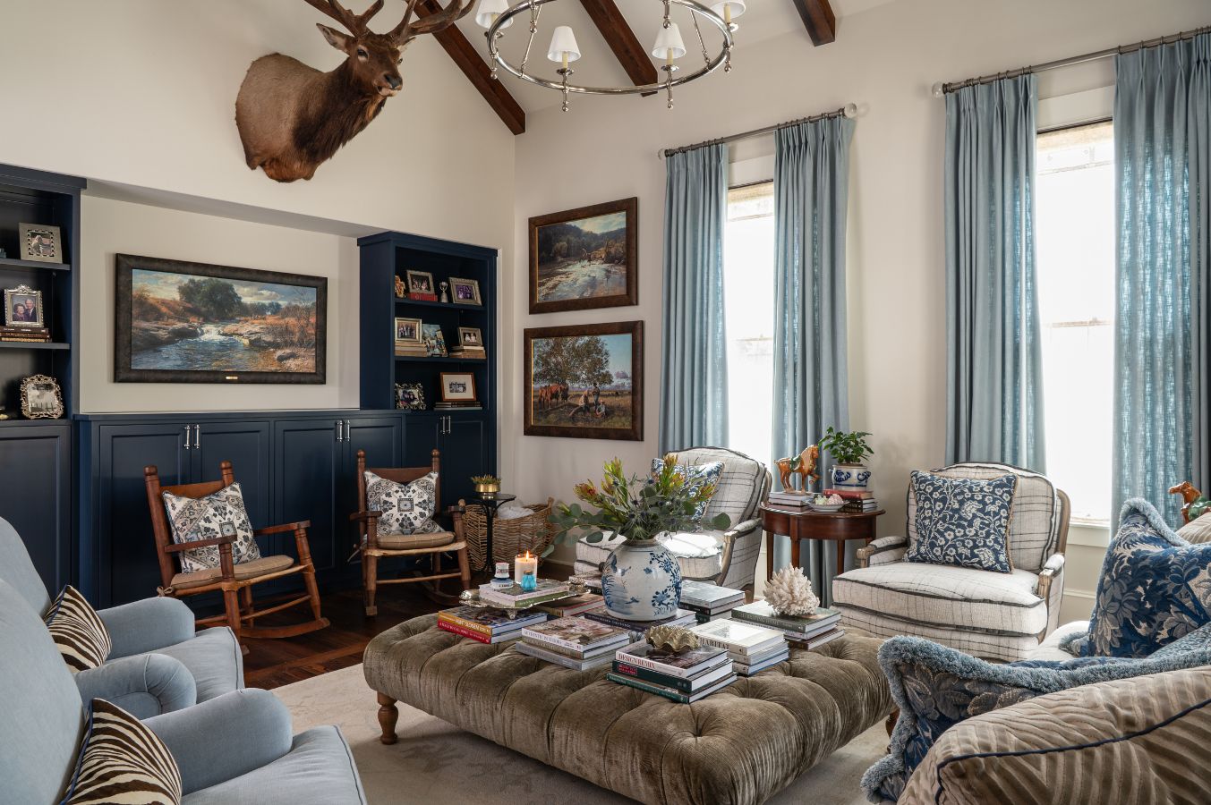 A cozy living room designed by an interior decorator in Boerne features blue cabinetry, rocking chairs, patterned curtains, comfy sofas, a large ottoman with books, landscape paintings, and a mounted deer head for classic Hill Country style.