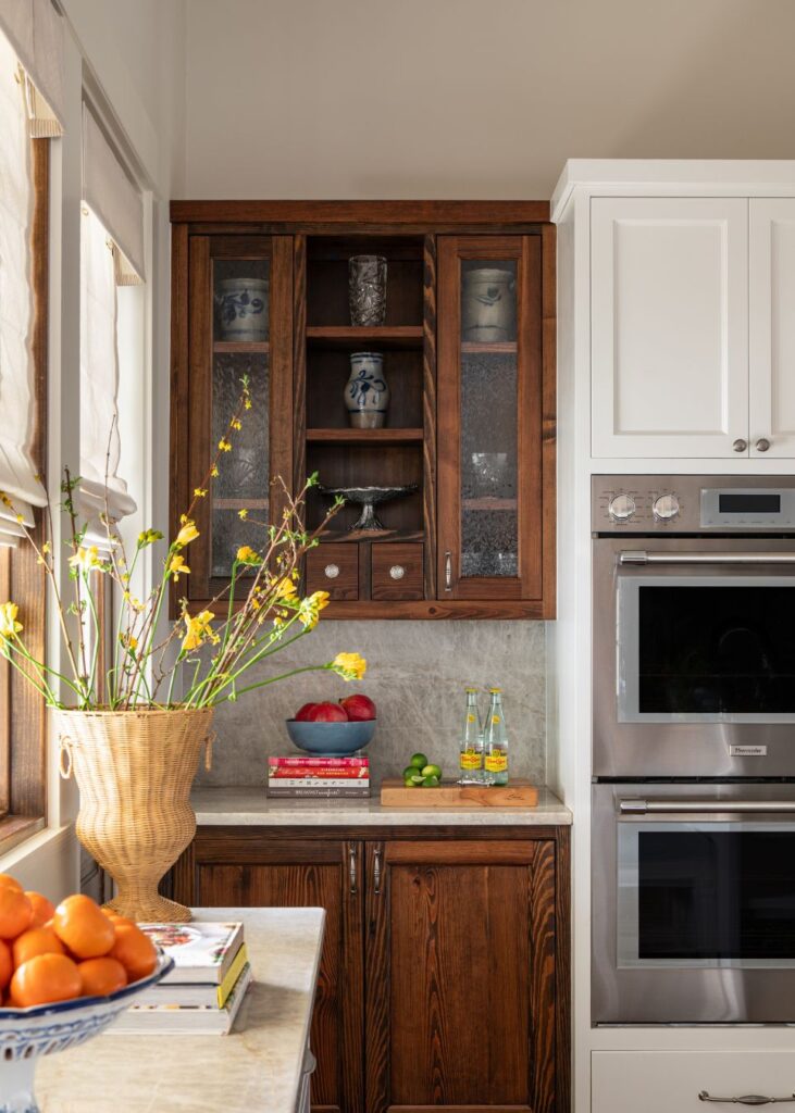 A kitchen corner by an interior design company in Boerne, TX, features dark wood lower cabinets, white uppers, stainless ovens, glass-front displays for ceramics, a vase with yellow flowers, stacked books, and fresh fruit on the counter.
