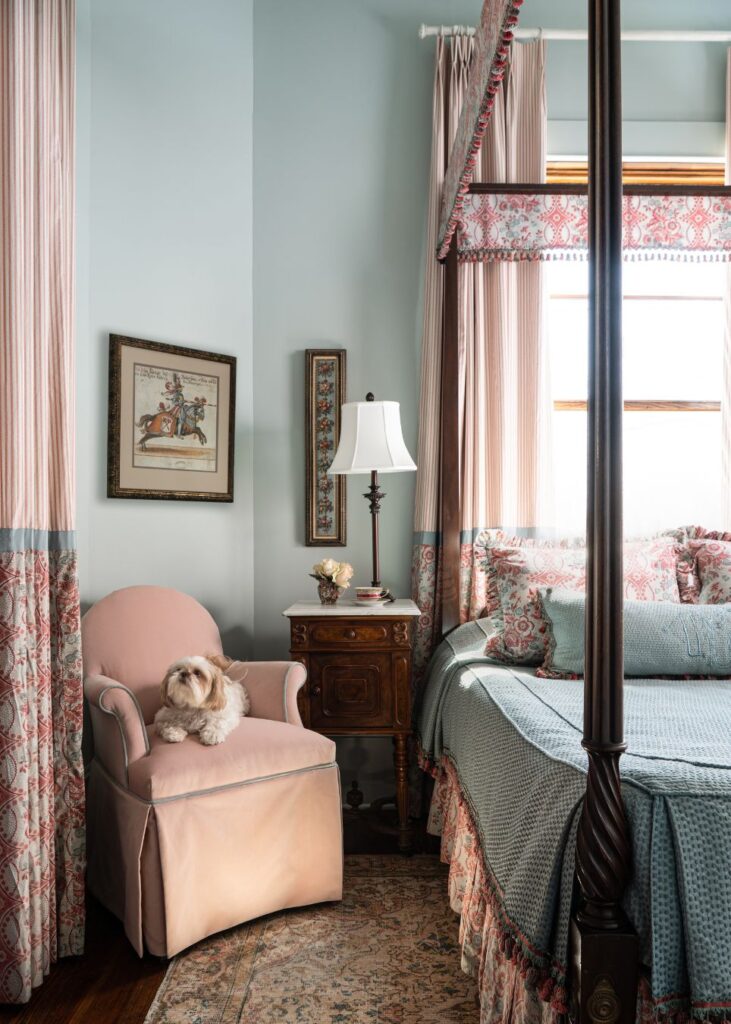 A small white dog sits on a pink armchair beside a wooden nightstand in a cozy, elegant bedroom styled by an interior designer Boerne TX, featuring patterned curtains, a four-poster bed, and soft pastel decor.