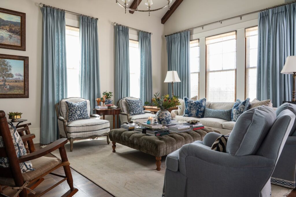 A cozy living room, styled by an interior designer Boerne TX, features large windows, light blue curtains, cushioned armchairs, a gray tufted ottoman, a beige rug, a sofa with blue pillows, wooden accents, books, and plants.
