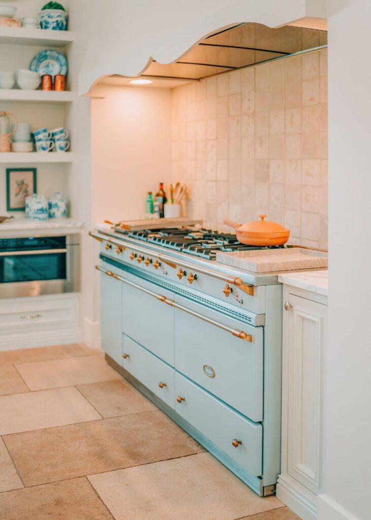 A pastel blue vintage-style stove with gold knobs is set against a tiled backsplash in a bright, elegant kitchen designed by an interior designer Boerne TX. An orange pot and open shelves with blue-and-white dishes add charm.