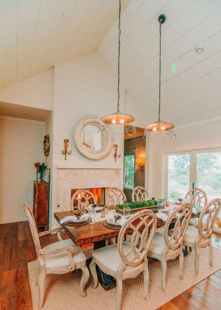 A cozy dining room styled by an interior designer in Boerne, TX, featuring a long wooden table for eight, elegant chairs, fireplace, round mirror, hanging lights, wood floors, and large windows that fill the space with natural light.