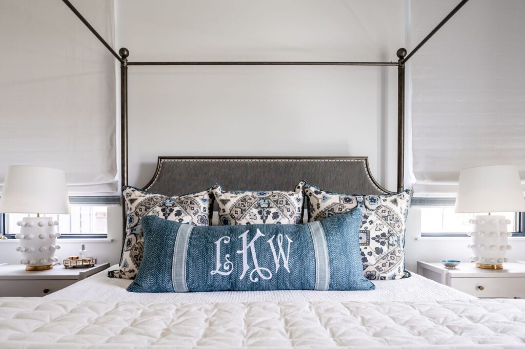 A neatly made bed with a navy and white patterned headboard, four patterned pillows, and a large blue monogrammed pillow showcases the touch of an expert interior decorator Boerne. Two white lamps with round bases sit on bedside tables on either side.