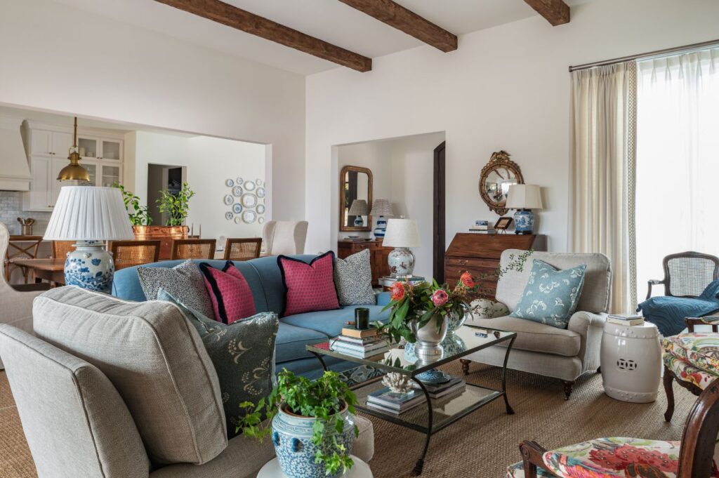 Cozy, well-lit living room with neutral sofas, colorful cushions, glass coffee table with books and flowers, potted plants, wooden beam ceiling, and vintage decor. Dining area and kitchen are visible in the background.