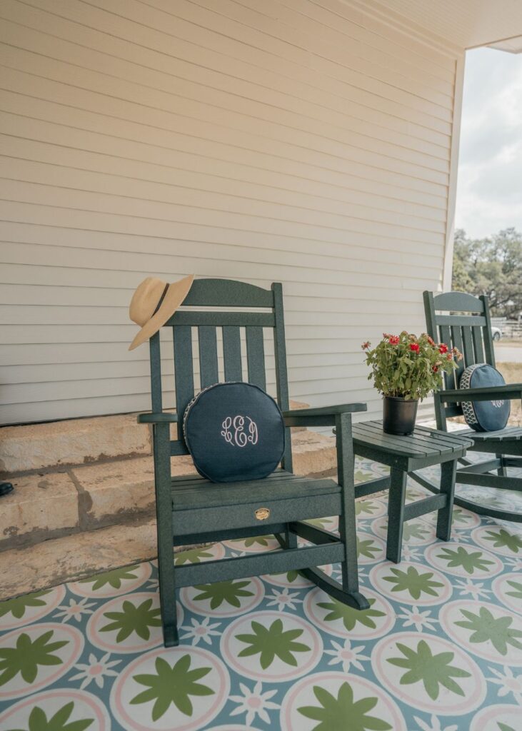 A green rocking chair with a round cushion and a straw hat rests on a patterned porch floor beside potted flowers—perfect inspiration for any interior designer Boerne TX seeking classic, welcoming touches in front of a light-colored house wall.