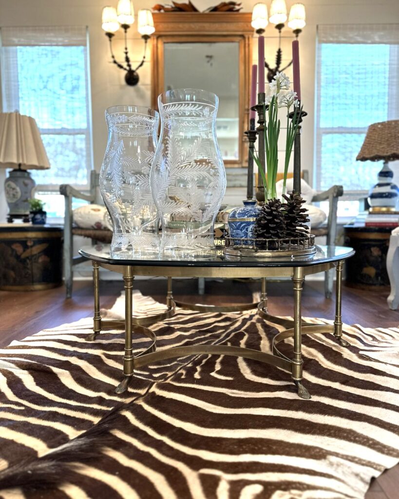 A living room styled by an interior design company in Boerne, TX, featuring a glass and brass coffee table on a zebra-print rug, two etched glass vases, candles, pinecones, vintage-style furniture, a large mirror, and bright windows.