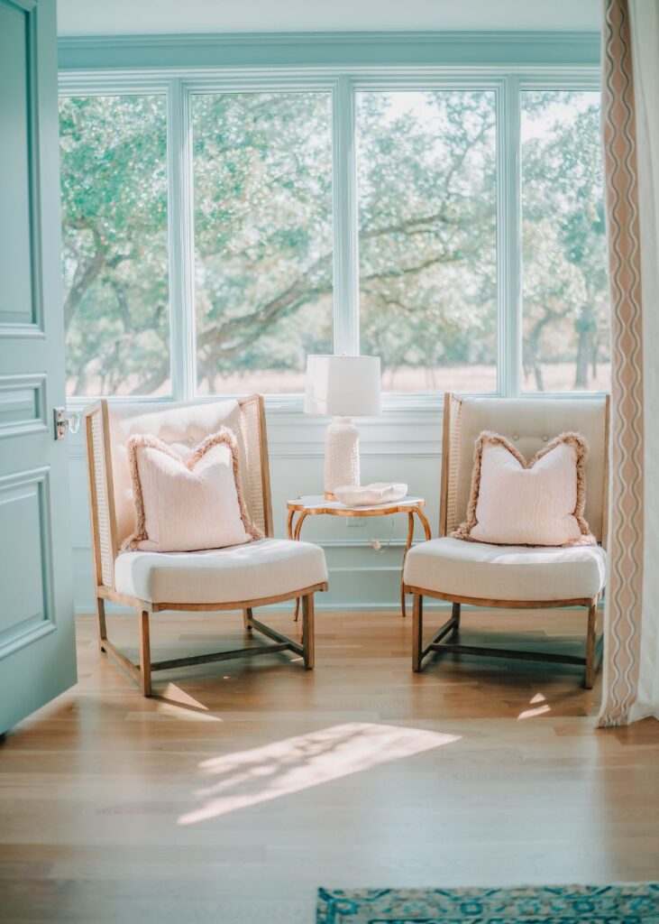 Two cushioned armchairs with pink throw pillows sit beside a small table with a white lamp and book. Large windows invite natural light, while an airy decor and light wood floor reflect the style of an experienced interior designer Boerne TX.