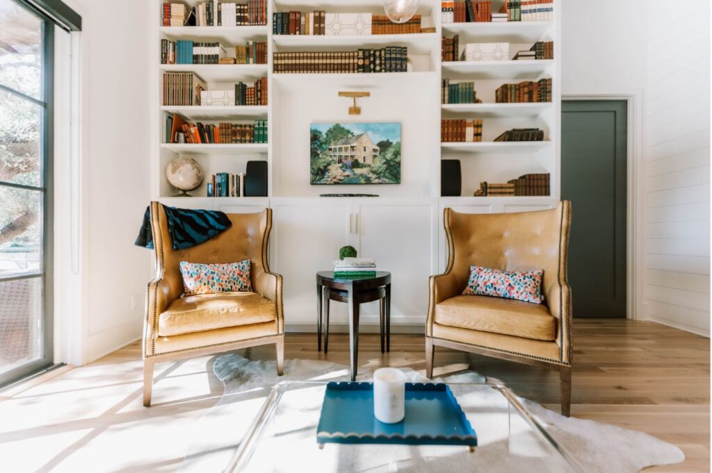 Two tan leather armchairs with colorful pillows face each other in a bright living room, styled by an interior design company Boerne TX. Bookshelves, decor, and art above the mantel complete this inviting space.