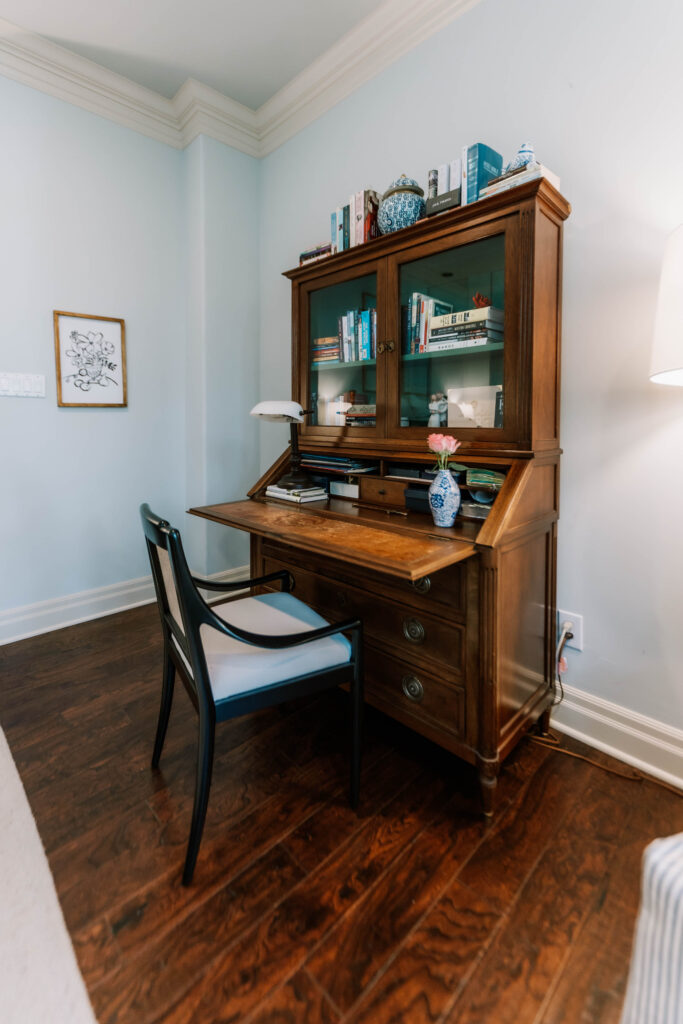 A vintage wooden desk with shelves and glass doors sits against a light blue wall—styled by an interior designer Boerne TX—with books, a lamp, and a vase arranged neatly. A black chair with a white cushion completes the charming look.