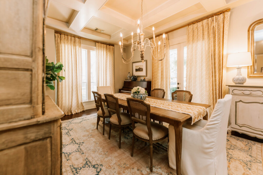 A warm, elegant dining room by an interior designer Boerne TX features a wooden table set for eight, beige upholstered chairs, a chandelier, large windows with flowing curtains, and classic decor including a mirror, plants, and a sideboard.