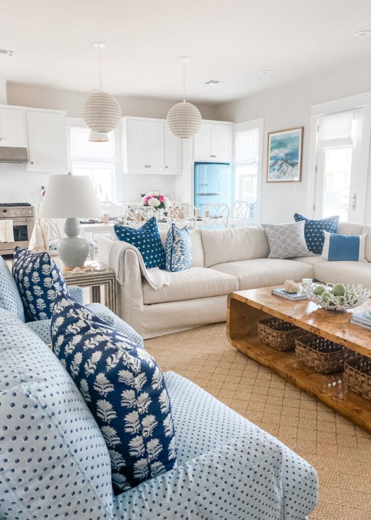 Bright, airy living room with coastal decor curated by an interior decorator Boerne, featuring white sofas with blue patterned pillows, a wooden coffee table, light blue armchair, woven rug, and a dining area with white chairs and a blue fridge in the background.