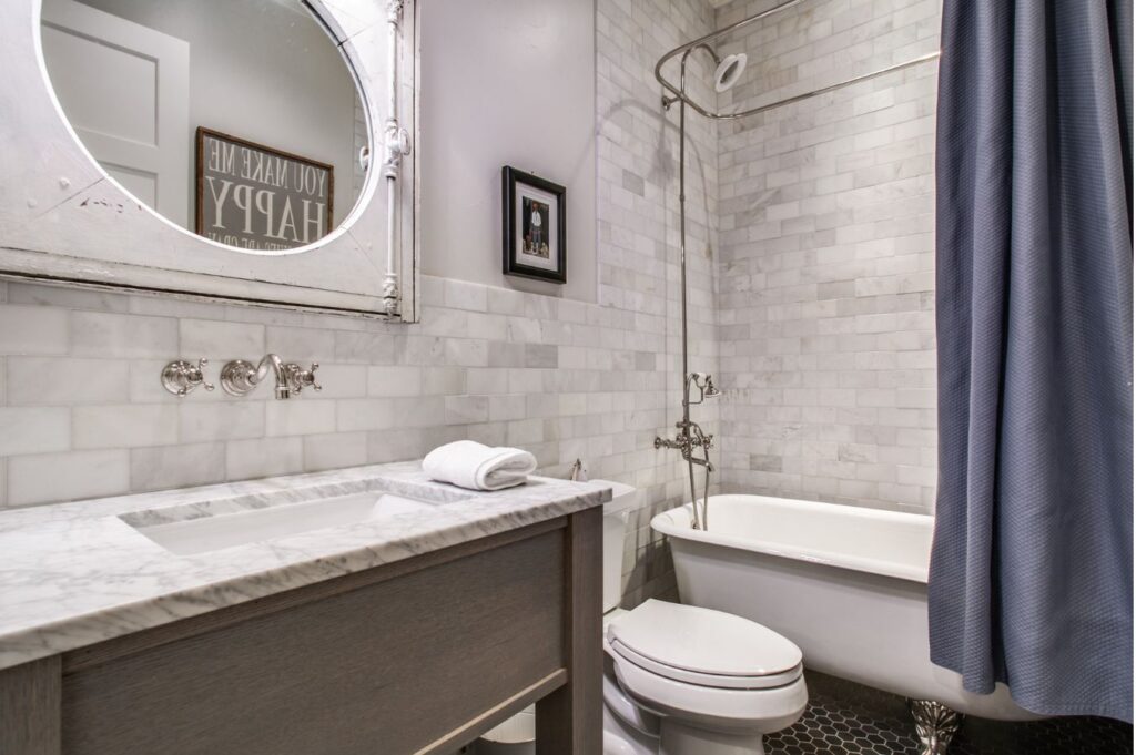 A bathroom designed by an interior decorator Boerne features a marble countertop, rectangular sink, wall-mounted faucet, and round mirror. A clawfoot tub with blue shower curtain sits beside the toilet and white subway tile walls.