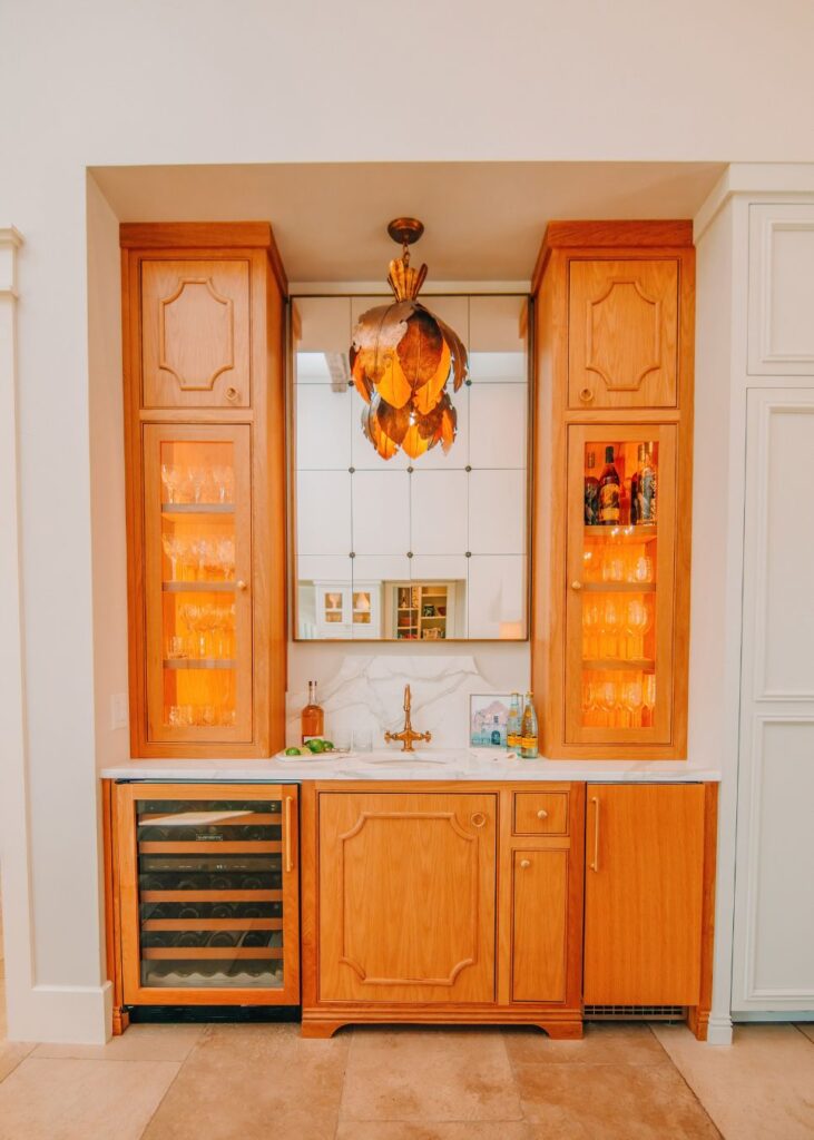 A built-in home bar with wooden cabinets, glass shelves displaying glassware, a wine fridge, brass faucet, and mirrored backsplash. Designed by an interior decorator Boerne, a decorative pendant light hangs above the countertop.