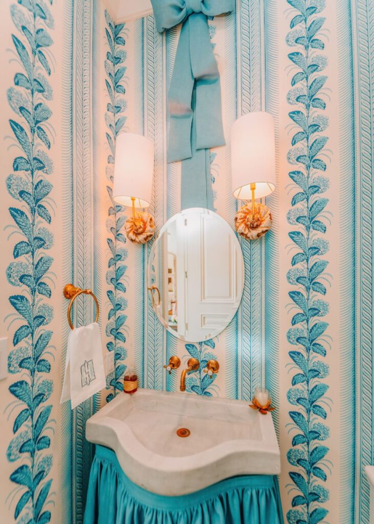 A small bathroom styled by an interior designer in Boerne, TX, features blue and white fern-patterned wallpaper, a round mirror, two wall lights, a wavy-edged sink with a blue skirt, and a large decorative blue bow above the mirror.